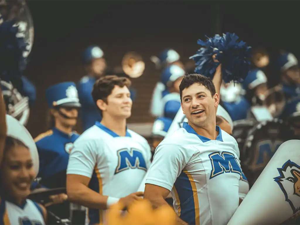 Cheerleaders and band members celebrate at a sports event, showcasing school spirit with vibrant uniforms and blue pom-poms.