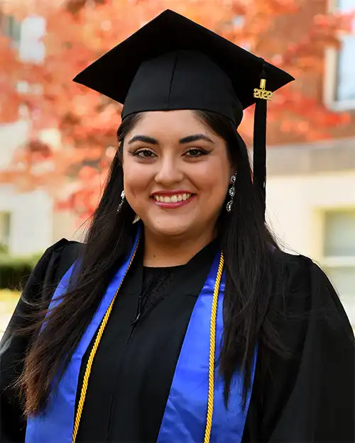 A smiling graduate in a black cap and gown with blue honors cords, standing outdoors against a background of autumn foliage.
