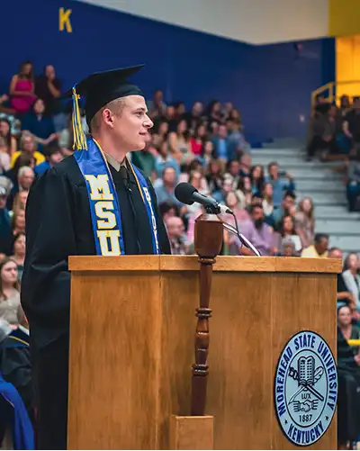 A graduate in cap and gown delivers a speech at a commencement ceremony, with a diverse audience in the background.