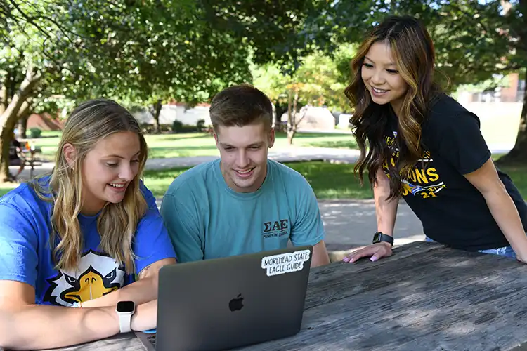 Three students sit outside on MSU campus with laptop.
