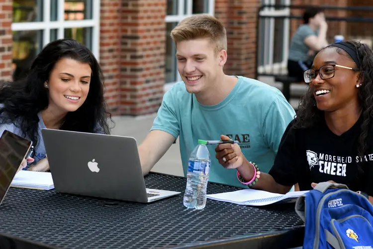 three students talking and laughing at a table around a laptop